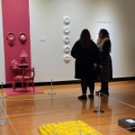 An art gallery. Closest to the viewer, there is an installation of yellow soap bars. On the wall, there are several round white artworks and a pink art installation. There are two figures are standing between the yellow and the pink installations talking. on February 27, 2026